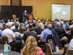 Photograph of a conference room with a speaker presenting at a podium and an audience seated facing a large screen displaying a presentation titled "2023 Leadership Summit." Attendees are dressed in business casual attire, with the room featuring neutral-colored walls, round tables, and chairs arranged to focus on the speaker and screen.