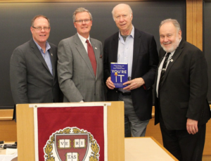 NPLI faculty members and co-authors of "You're It: Crisis, Change, and How to Lead When It Matters Most," Joe Henderson, Eric McNulty, David Gergen, and Lenny Marcus. They are standing together at the front of a classroom behind a podium with the Harvard shield. Joe Henderson is holding a copy of the book. 